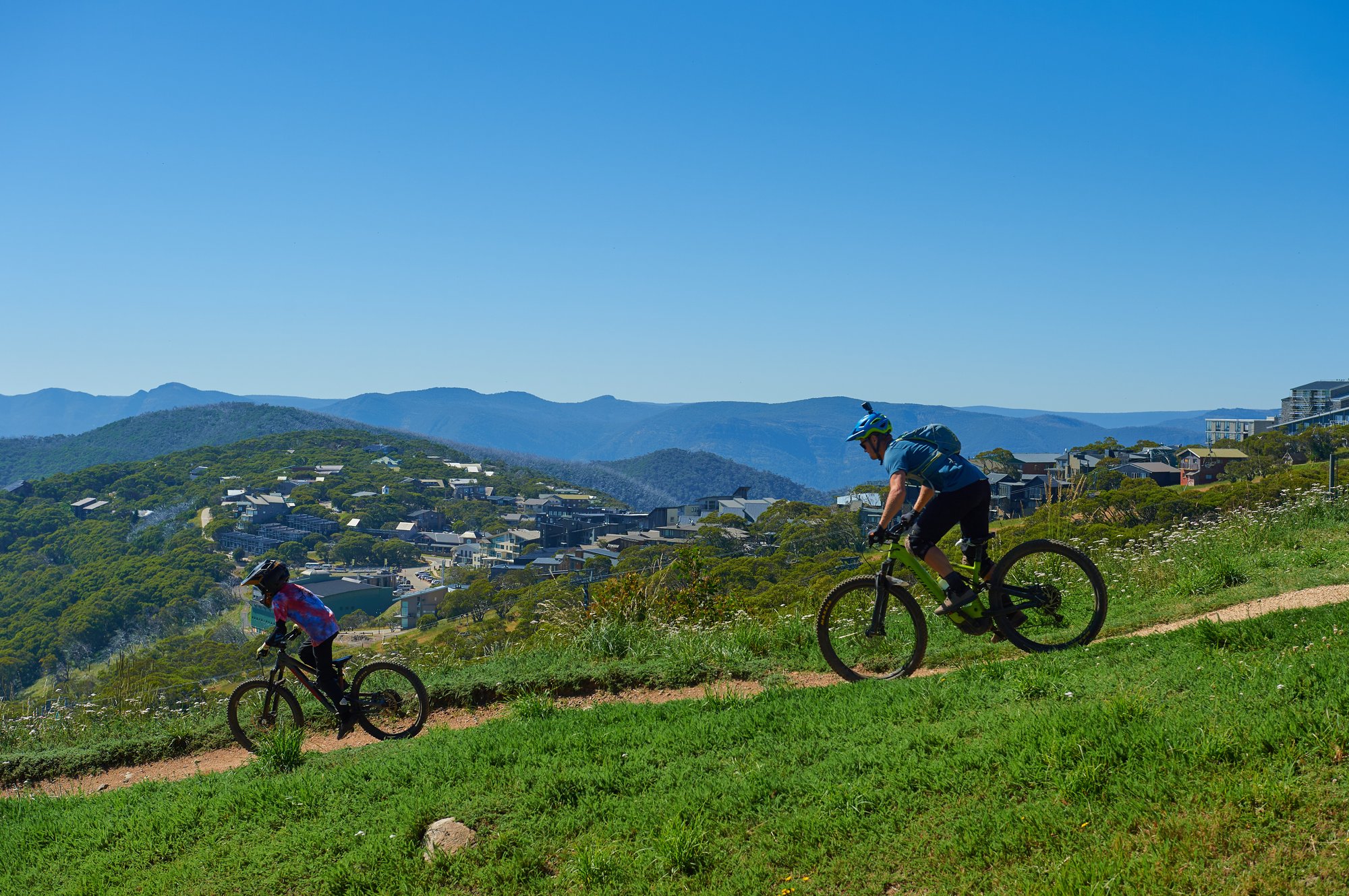 Two mountain bikers riding a single trail with the Mt Buller village and sweeping mountain views behind them