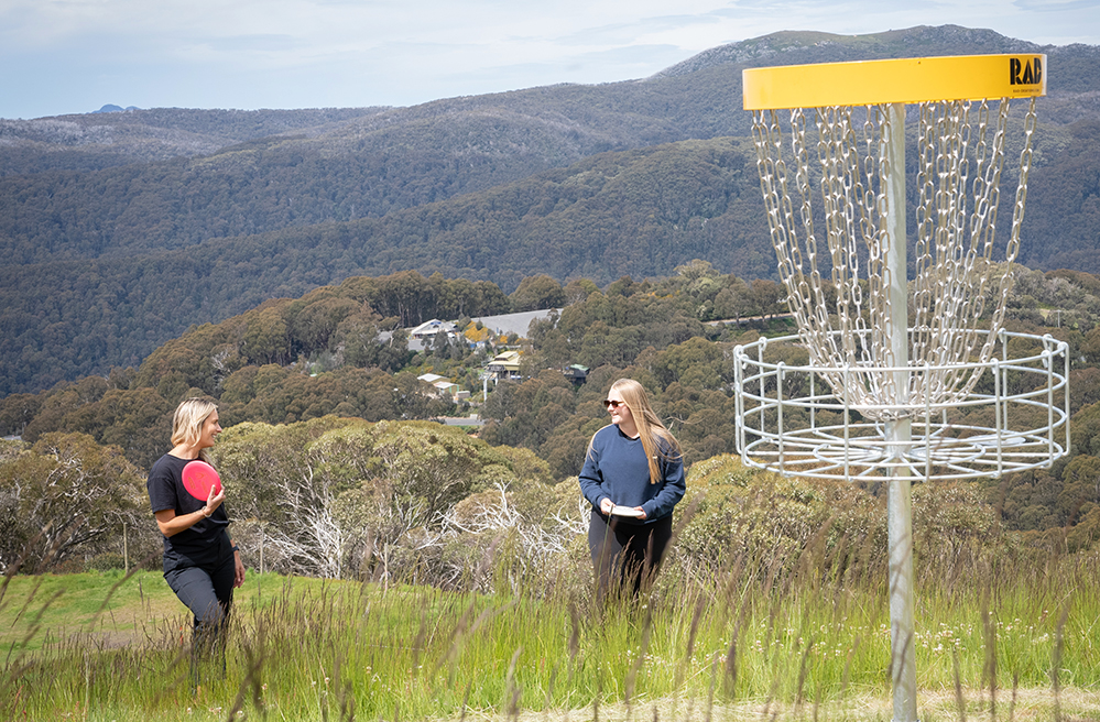 Friends play disc golf on Mt Buller on a sunny summer day walking through lush green grass with sweeping alpine views to nearby Mt Stirling
